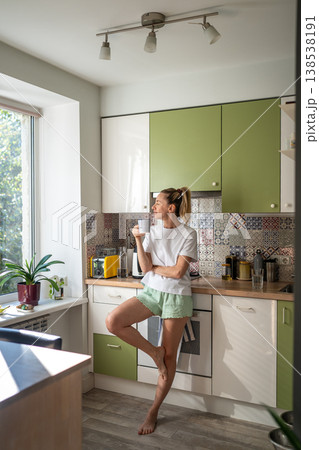 Cheerful woman standing in kitchen looking at window and enjoying morning coffee on weekend. Cheerful woman standing in kitchen looking at window and enjoying morning coffee on weekend. 138538191