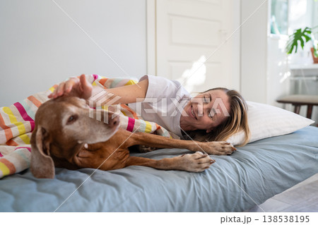 Smiling relaxed woman cuddling with Vizsla dog in bed in morning, sharing joyful moments with pet. Smiling relaxed woman cuddling with Vizsla dog in bed in morning, sharing joyful moments with pet. 138538195