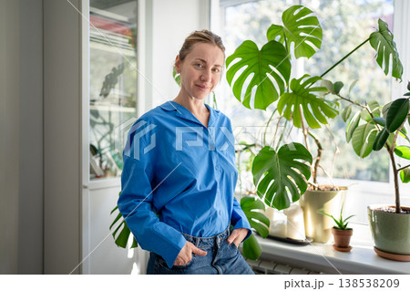 Cheerful happy middle aged woman enjoying indoor gardening, posing near potted plants at windowsill. 138538209