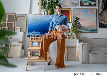 Smiling successful female painter posing in front of artwork on easel in modern art home studio. 138538212