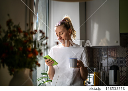 Relaxed thoughtful woman drinking coffee scrolling smartphone at cozy sunny kitchen beside window. 138538258