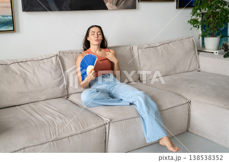 Woman waving paper fan cooling herself during summer heatwave, feeling stress and dehydration. Woman waving paper fan cooling herself during summer heatwave, feeling stress and dehydration. 138538352