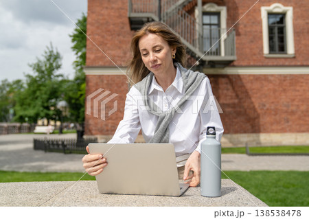 Cheerful businesswoman working on laptop outside in park, preparing for online business conference. Cheerful businesswoman working on laptop outside in park, preparing for online business conference. 138538478