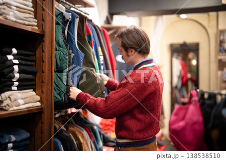 Mindful man customer picking clothing in second hand vintage boutique, choosing different vests. 138538510