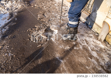 Street cleaner breaking ice with breaker tool, cleaning iced formation from drainpipe on sidewalk. Street cleaner breaking ice with breaker tool, cleaning iced formation from drainpipe on sidewalk. 138538562