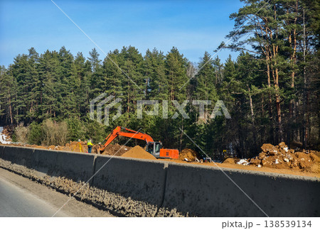 Orange excavator moving earth to form highway embankment while surveyor with total station measures site beside pine forest under clear blue sky during daytime construction. Orange excavator moving earth to form highway embankment while surveyor with total station measures site beside pine forest under clear blue sky during daytime construction. 138539134