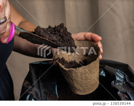 Woman's hands filling an organic peat pot with dark potting soil using a garden trowel, preparing for seedling transplantation and advocating for sustainable gardening practices. 138539144