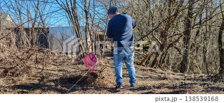 Man wearing jeans and a blue sweater raking dry leaves and branches in a neglected backyard during a sunny early spring day, engaging in garden cleanup and yard work. 138539168