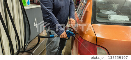 A man fills up an orange car at a gas station, holding a blue nozzle in the tank. City night scene showing gas mileage, cost and everyday travel expenses. 138539197