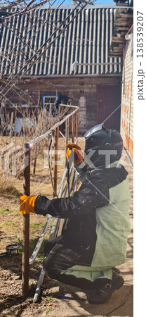 Welder wearing protective helmet and gloves performing arc welding on a metal fence post, creating bright sparks, demonstrating manual labor and repair work in a rural setting. 138539207