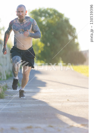 Muscular tattooed man running outdoors on path during sunrise Muscular tattooed man running outdoors on path during sunrise 138539396