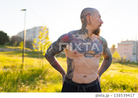 Tattooed man standing shirtless in a park at sunset 138539417
