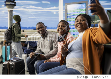 Family of three taking pictures on phone in bright airport waiting area, parents and teenage daughter on a bench with plane ticket and travel documents ready. Boarding flight in cheerful good mood. Family of three taking pictures on phone in bright airport waiting area, parents and teenage daughter on a bench with plane ticket and travel documents ready. Boarding flight in cheerful good mood. 138539737