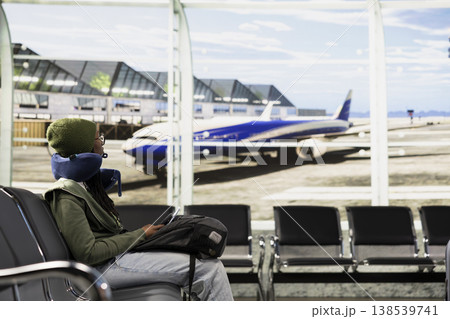 African american solo traveler sitting in airport lounge and looking at runway, waiting for departure and boarding international flight. Woman relaxing by the terminal gate with her backpack. African american solo traveler sitting in airport lounge and looking at runway, waiting for departure and boarding international flight. Woman relaxing by the terminal gate with her backpack. 138539741
