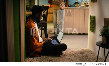 African american person, relaxing with laptop, sitting on the floor at home, browsing websites and enjoying free time on the weekend. Comfortable domestic life with tranquility and gadgets. African american person, relaxing with laptop, sitting on the floor at home, browsing websites and enjoying free time on the weekend. Comfortable domestic life with tranquility and gadgets. 138539871