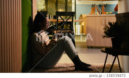 African American woman relaxing alone at home and checking messages, scrolling social media on smartphone. Comfortable domestic setting highlighting calm lifestyle, everyday relaxation. African American woman relaxing alone at home and checking messages, scrolling social media on smartphone. Comfortable domestic setting highlighting calm lifestyle, everyday relaxation. 138539872