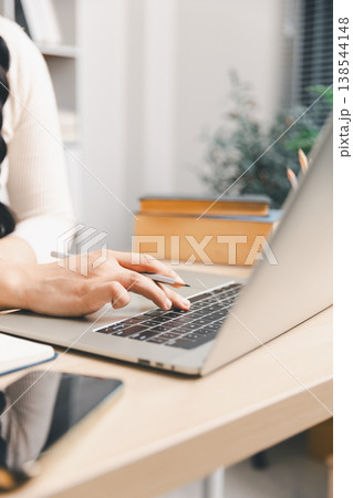 Close up of young Asian woman using laptop and taking notes at home desk with smartphone nearby. Remote study, online learning, digital research, planning and daily productivity concept. Close up of young Asian woman using laptop and taking notes at home desk with smartphone nearby. Remote study, online learning, digital research, planning and daily productivity concept. 138544148