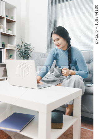 Asian woman using laptop while holding coffee mug on sofa at home, showing remote work, online productivity and cozy home office lifestyle in a bright modern living room interior. Asian woman using laptop while holding coffee mug on sofa at home, showing remote work, online productivity and cozy home office lifestyle in a bright modern living room interior. 138544180