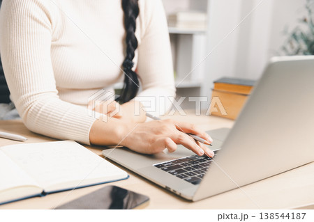 Close up of young Asian woman using laptop and taking notes at home desk with smartphone nearby. Remote study, online learning, digital research, planning and daily productivity concept. 138544187