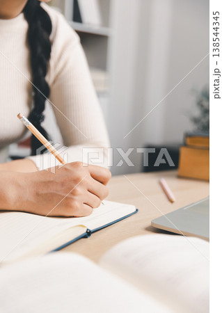 Close up of woman writing notes in notebook beside laptop and open book at home desk. Daily study routine, online learning, homework, remote education and focused productivity concept. 138544345