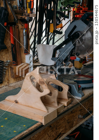 Carpentry workbench. Tool storage. Electric miter saw with safety wooden push block on table with hanging equipment collection in garage interior. 138544675