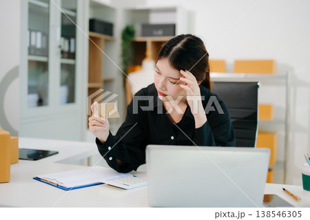 Stressed Asian woman sitting at office desk with parcels and laptop, overwhelmed by online business problems, workload pressure 138546305