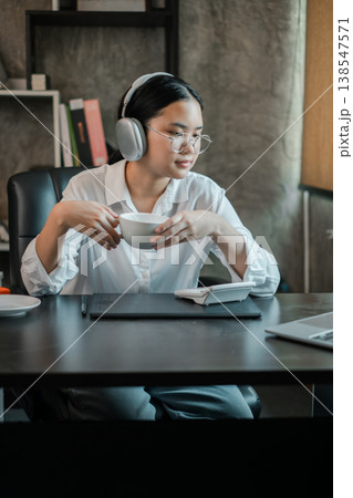 Woman in office wearing headphones, holding coffee, and using a tablet. Modern workspace with relaxed atmosphere. 138547571
