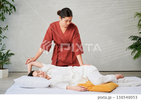 Masseuse making thai yoga massage. Woman therapist doing traditional massage treatment, stretching female patient's body. Client lying on mat, enjoying hand massage in calm atmosphere. 138548053