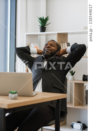 An African American man in a suit leans back in his chair with his hands behind his head, smiling contentedly while working on his laptop An African American man in a suit leans back in his chair with his hands behind his head, smiling contentedly while working on his laptop 138549017