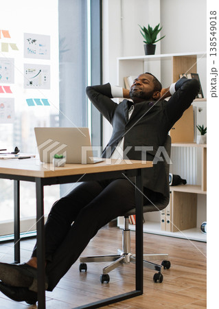 An African American man in a suit leans back in his office chair with his hands behind his head, eyes closed in a moment of relaxation and satisfaction An African American man in a suit leans back in his office chair with his hands behind his head, eyes closed in a moment of relaxation and satisfaction 138549018