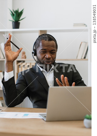 An African American man wearing a headset and suit is actively engaged in a video conference call on his laptop, gesturing with his hands An African American man wearing a headset and suit is actively engaged in a video conference call on his laptop, gesturing with his hands 138549031