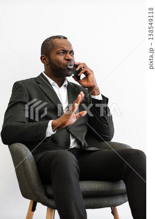An African American man in a suit sits in a chair while talking on his phone with a frustrated expression and gesturing with his hand 138549118
