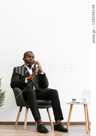 An African American man in a suit sits in a chair, engaged in a phone conversation with a serious expression, gesturing with his hands An African American man in a suit sits in a chair, engaged in a phone conversation with a serious expression, gesturing with his hands 138549144