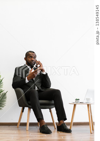 An African American businessman in a suit sits in a chair while talking on his mobile phone, looking thoughtful An African American businessman in a suit sits in a chair while talking on his mobile phone, looking thoughtful 138549145