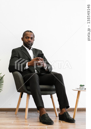 An African American man in a suit sits in a chair holding a smartphone and looking at the camera An African American man in a suit sits in a chair holding a smartphone and looking at the camera 138549146
