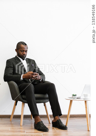 An African American man in a suit sits in a chair while looking at his smartphone, with a laptop and coffee cup on a nearby table 138549166