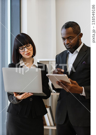 A woman and an African American man in business attire collaborate using a laptop and tablet in an office setting 138549186