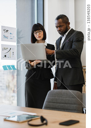A focused African American man and a woman in business attire collaborate over a laptop, reviewing charts and data on a glass wall A focused African American man and a woman in business attire collaborate over a laptop, reviewing charts and data on a glass wall 138549203