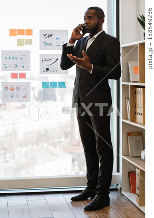 An African American man in a suit talks on his phone while gesturing with his hand near a window with charts and sticky notes An African American man in a suit talks on his phone while gesturing with his hand near a window with charts and sticky notes 138549236