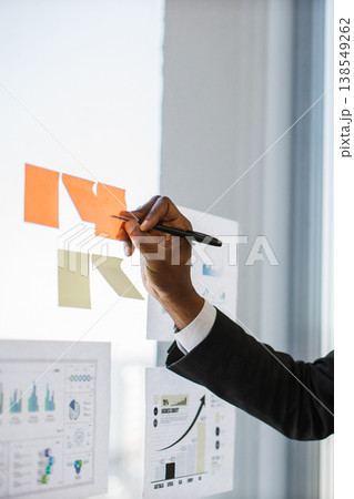 An African American hand in a suit writes on sticky notes attached to a glass wall with charts and graphs in the background An African American hand in a suit writes on sticky notes attached to a glass wall with charts and graphs in the background 138549262