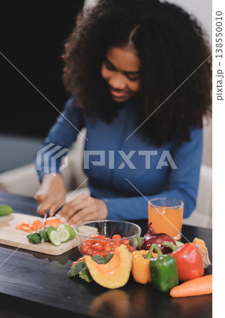 Young nutritionist showing bell peppers during an online video call consulting about healthy eating, sitting at home at a table with vegetables, laptop and measuring tape 138550010