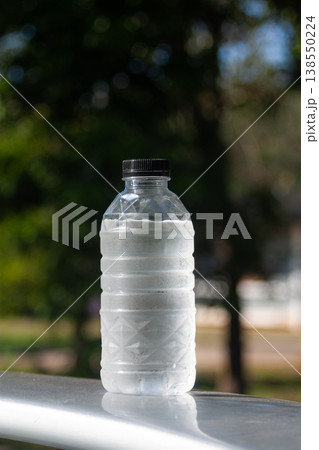 A cold plastic water bottle with condensation stands outdoors, catching sunlight against green blur. A cold plastic water bottle with condensation stands outdoors, catching sunlight against green blur. 138550224
