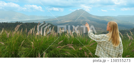 A woman stands in grassy fields while pointing at a volcano 138555273