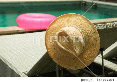 A straw hat rests on a chair near a pool with a float A straw hat rests on a chair near a pool with a float 138555304