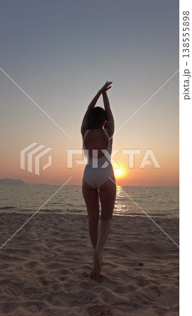Woman in white swimsuit walking on beach to sunset over sea 138555898