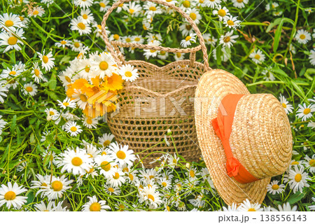 A straw hat, wicker bag with bouquet of white and yellow daisy flowers on a blooming meadow A straw hat, wicker bag with bouquet of white and yellow daisy flowers on a blooming meadow 138556143