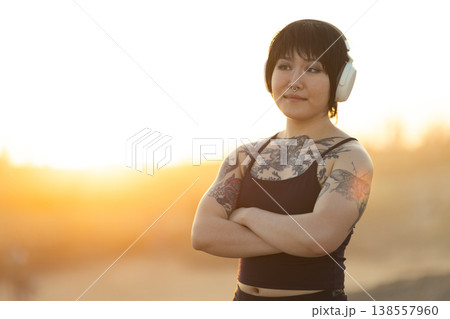 Tattooed confident woman wearing wireless headphones at sunset Tattooed confident woman wearing wireless headphones at sunset 138557960