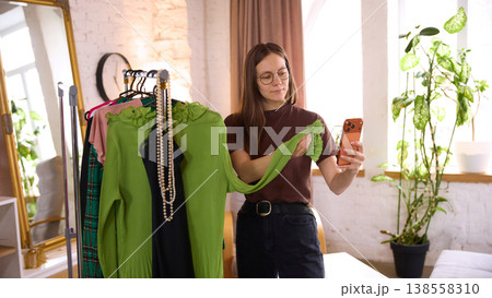 Woman holding green garment and smartphone comparing clothes before purchase 138558310
