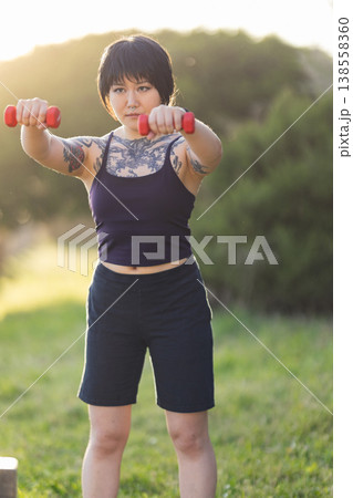 Tattooed young woman exercising with dumbbells outdoors 138558360