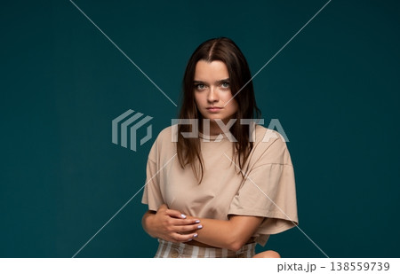 Woman with dark hair and a focused expression sitting, hands clasped, wearing a casual t-shirt against a teal backdrop 138559739